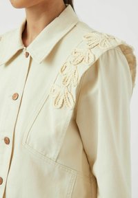 Chaqueta beige claro con un gran collar, cierre frontal de botones y bordado decorativo en color crema a lo largo del hombro.