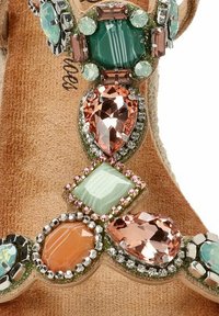 Tirante decorativo de sandalia que presenta piedras preciosas mezcladas en tonos verde azulado, rosa y melocotón, sobre una base texturizada de color beige claro con acentos metálicos.