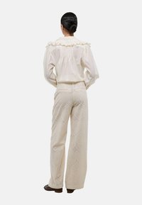 Blouse créme à volants avec des plis à l'arrière, assortie à un pantalon texturé taille haute avec un motif géométrique. Chaussures décoratives sombres.