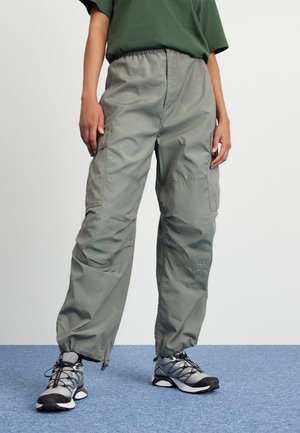 Persoon die losse grijze cargobroek en grijze zwarte vetersneakers draagt, staand op een blauw tapijt.