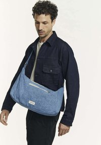 Sac en tissu bleu avec une poche zippée diagonale. Conçu dans une forme décontractée. Porté par une personne en veste en denim foncé et pantalons noirs.