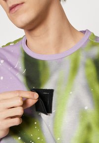 Flerfärgad kortärmad skjorta med lila, grön och vit mönstring; har en svart kardborrelapp nära halsen. Mjuk tygkänsla.