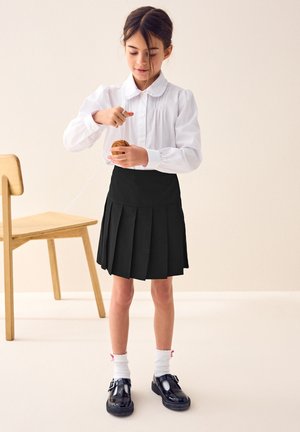 Jong meisje in witte blouse en zwarte rok speelt met een houten jojo naast een houten stoel op een lichte achtergrond.