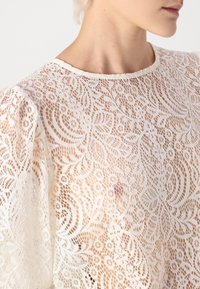 Blouse en dentelle blanche avec des motifs floraux complexes, texture transparente, col rond et manches bouffantes. Design délicat et léger.