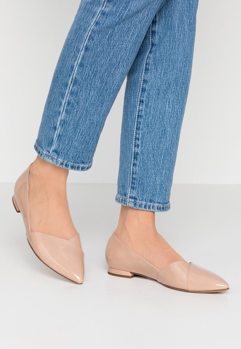 Des pieds portant des ballerines nude en cuir verni à bout pointu, associés à un jean en denim bleu, contre un fond clair et uni.