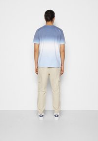 Camiseta de fondo azul claro con gradiente y mangas cortas, combinada con pantalones cargo beige. Mostrando una textura suave y un ajuste relajado desde la vista trasera.