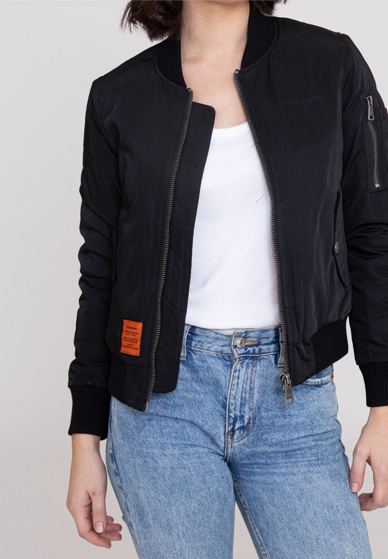 Personne portant un blouson bomber noir zippé sur un débardeur blanc côtelé et un jean taille haute bleu clair, debout sur un fond uni.