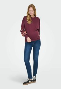 Maglione borgogna con maniche a fantasia floreale, abbinato a jeans skinny blu e sneakers nere con dettagli bianchi e beige.