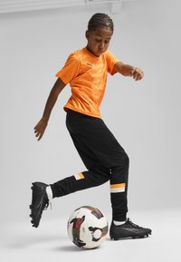 Oranža īsām piedurknēm krekls ar teksturētu rakstu; melnas bikses ar oranžiem un baltiem akcentiem; melni futbola zābaki; balts futbols ar rakstu.