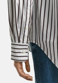 Camicia a righe con linee verticali bianche e marroni scure, polsini con bottoni, tessuto di cotone liscio e taschino sulla manica sinistra. Vestibilità casual.