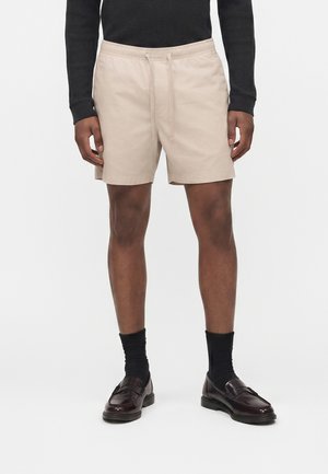 Άνδρας που φοράει μπεζ σορτς με κορδόνι, μαύρο μακρυμάνικο μπλουζάκι, μαύρες κάλτσες και σκουρόχρωμα καφέ δερμάτινα loafers, στέκεται μπροστά σε απλό φόντο.