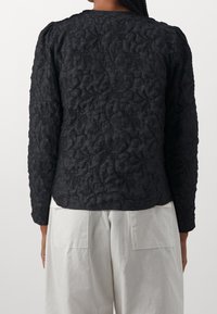 Maglietta nera a maniche lunghe con texture e design trapuntato, caratterizzata da un collo rotondo e un orlo leggermente curvo, abbinata a pantaloni di colore chiaro.