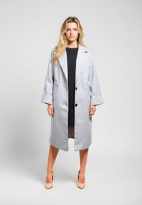 Gabardina impermeable gris claro con frente abierto, solapas grandes con muesca, dos botones y bolsillos laterales. Usada sobre un vestido negro.