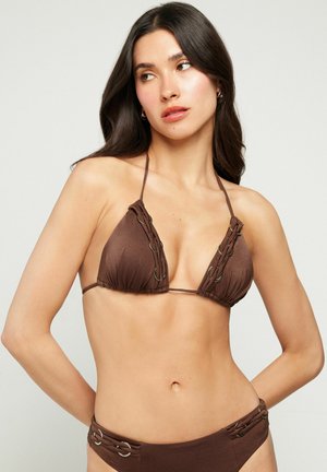 Femme aux longs cheveux foncés portant un bikini marron avec des anneaux métalliques, posant devant un fond clair uni.