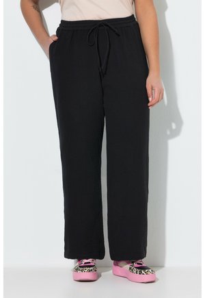 Person iført sorte bukser med snøre og pink leopardmønstrede sneakers, stående mod en hvid væg med den ene hånd i lommen.