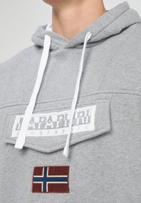 Sudadera con capucha gris hecha de tejido suave. Cuenta con un bolsillo frontal con una etiqueta blanca y un parche bordado de la bandera noruega. Cuerdas blancas.