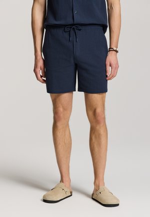 Mann trägt marineblaue Shorts mit Kordelzug, passendes Hemd, beige Slip-On-Schuhe und Armbänder am linken Handgelenk, steht vor neutralem Hintergrund.