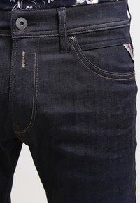 Mörka denimjeans med en slät yta, kontrasterande sömmar och en metallknapp i midjan. Har en sidficka och varumärkesetikett.