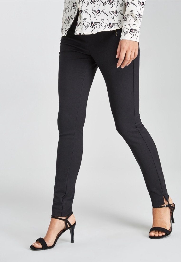 Pantalons noirs ajustés avec des fentes sur les chevilles, en matière lisse, associés à des sandales à talons hauts noires et à un chemisier à motifs.