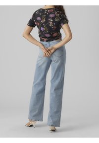 Haut court à manches courtes à fleurs noires associé à un jean denim bleu clair taille haute. Le modèle porte des sandales à talons beige. Vue de dos.