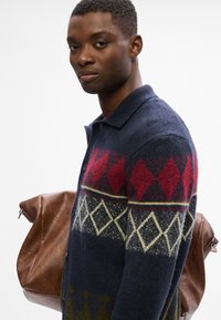 Granatowy sweter z splotu z czerwonymi i zielonymi wzorami w diamenty, z kołnierzykiem i guzikami, w połączeniu z brązową skórzaną torbą.