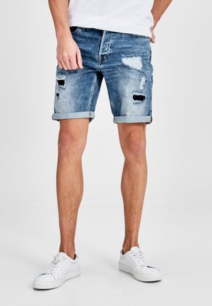 Hellblaue Jeansshorts mit Used-Look, einschließlich aufgerissener Stellen und umgeschlagenen Säumen. Kombiniert mit weißen Turnschuhen.