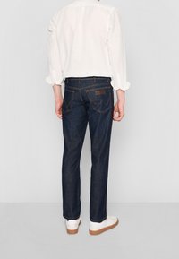 Mörkblå jeans med rak passform, fem fickor, diskret sömnad och en märkespäls på baksidan av midjebandet. Vit skjorta och sneakers.