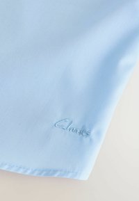 Hele sinise kangas sujuva tekstuuriga, millel on alumises paremas nurgas samas sinises niidis brodatuur "Clarks" logo.