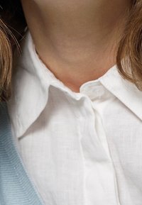 Bílá košile na knoflíky s texturovanou látkou, s špičatým límcem a viditelnými knoflíky, vrstvená pod světle modrým svetrem.
