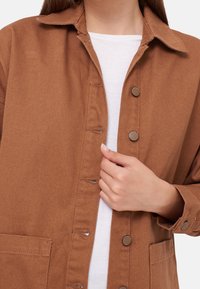 Hnědá bunda na knoflíky s límcem, dvěma předními kapsami a texturovanou látkou. Nositá přes bílou tričko.