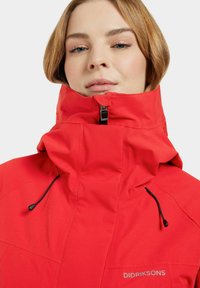 Veste imperméable rouge avec col montant, fermeture éclair et cordons de serrage réglables. Présente des détails de couture et un logo sur la poitrine.
