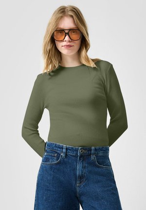 Jonge vrouw met blond haar, met oranje getinte aviator-zonnebril, olijfgroene geribbelde trui en een high-waisted blauwe jeans met een hartvormige knoop.