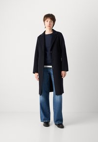 Manteau en laine bleu marine avec un col cranté, devant double boutonnage et manches longues, porté sur un haut ajusté bleu marine et un jean à jambes larges.