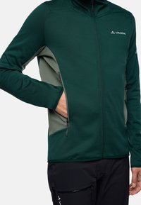 Giacca softshell verde con accenti grigi, tasche con zip, colletto alto e tessuto strutturato; presenta una zip frontale e un logo sul petto.