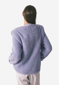 Futrzana fioletowa kurtka polarowa z okrągłym dekoltem i opuszczonymi ramionami, z fakturowaną powierzchnią i luźnym krojem, noszona na paskowanych spodniach.