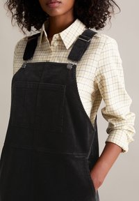 Salopette en velours noir avec bretelles ajustables et poche à l'avant, portée par-dessus une chemise à carreaux jaunes et blancs avec les manches retroussées.