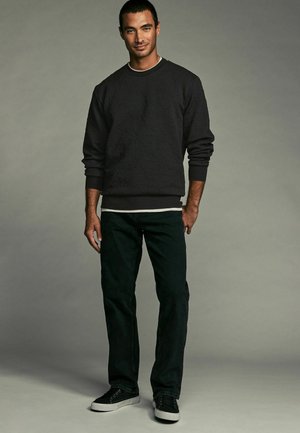 Sudadera negra texturizada con puños acanalados, llevada sobre una camisa blanca. Combinada con jeans oscuros y zapatillas negras, el modelo está de pie sobre un fondo gris.