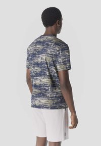 Mann mit kurzem lockigem Haar, der ein marineblaues und beiges gemustertes T-Shirt sowie hellfarbene, strukturiert wirkende Shorts trägt und mit dem Rücken zur Kamera steht.