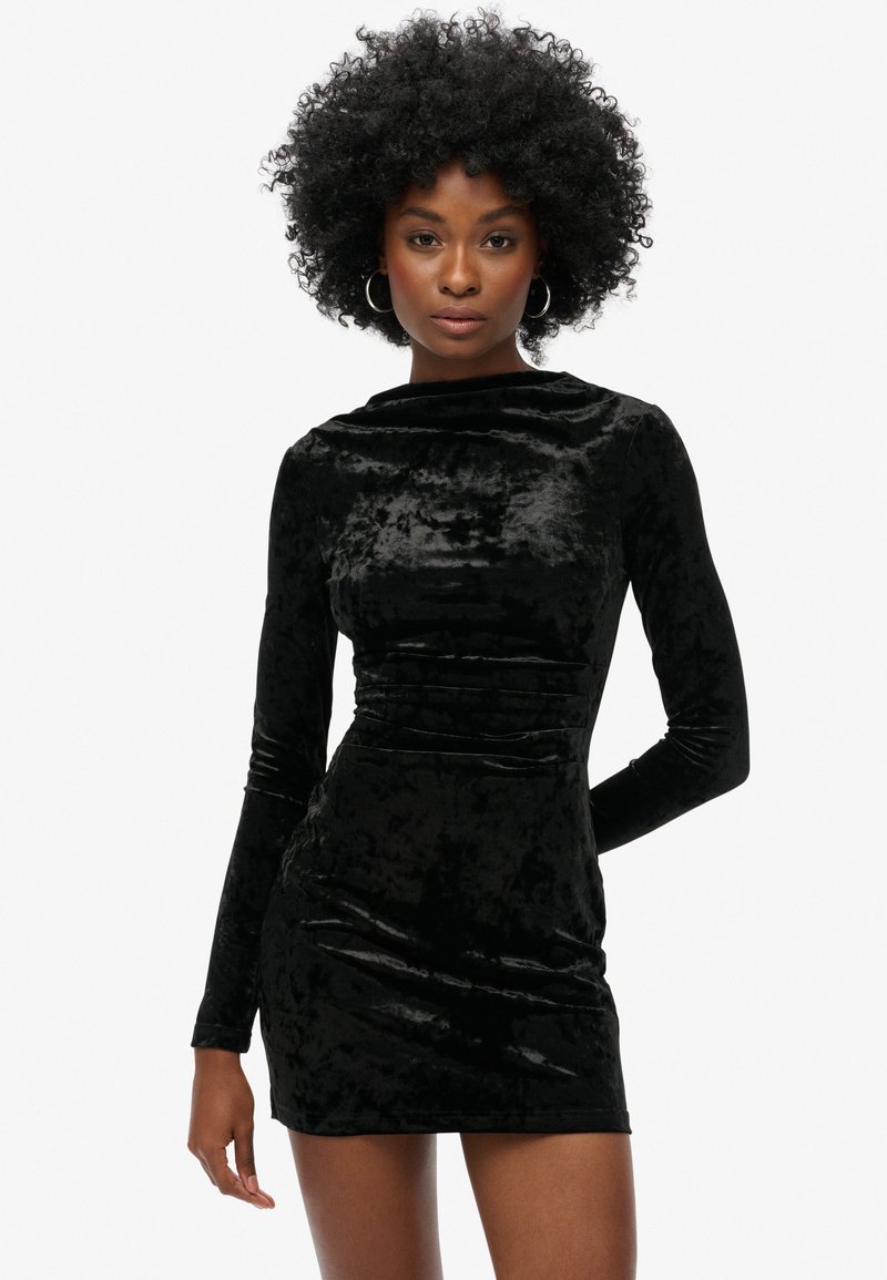 Vestido de terciopelo negro de manga larga con forma ajustada, cuello redondo y sutiles detalles de fruncido, que presenta una textura suave y lisa.