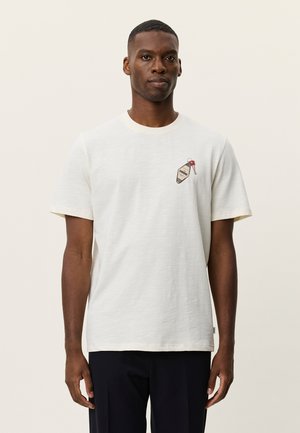 Άνδρας που φοράει απλό λευκό κοντομάνικο μπλουζάκι με ένα μικρό γραφικό ενός κλειδιού και ταυτότητας στην αριστερή πλευρά του στήθους, σε συνδυασμό με σκούρο παντελόνι.