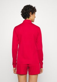 Rotes Sportoberteil mit hohem Kragen und langen Ärmeln, kombiniert mit passenden roten Shorts. Glatte Textur, enge Passform und keine sichtbaren Muster.