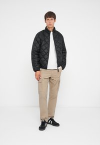 Giacca bomber nera trapuntata con colletto alto, abbinata a una maglietta bianca, pantaloni cargo beige e sneaker nere.