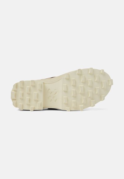 Semelle de chaussure avec une surface en caoutchouc texturé de couleur crème présentant de multiples crampons pour l'adhérence et la stabilité. Détail du logo central présent.