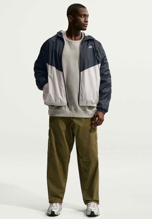Hombre con cortavientos Nike azul marino y blanco, sudadera gris, pantalones cargo verde oliva y zapatillas blancas, de pie con las manos en los bolsillos.