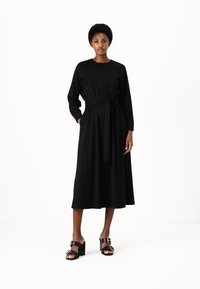 Svart långärmad klänning med en figurnära bodice, ett åtdraget midjeband och en svävande A-linjeformad kjol. Kombinerad med blockklackade svarta sandaler.