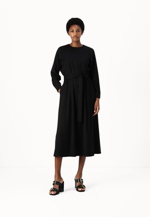Robe noire à manches longues avec un corsage ajusté, une ceinture à la taille cintrée et une jupe évasée fluide. Associée à des sandales noires à talons blocs.