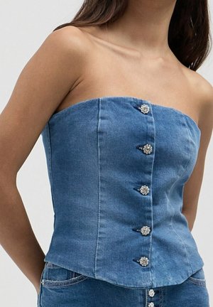 Kvinde iført en blå denim korsettop uden stropper med dekorative sølvblomsterknapper foran, kombineret med blå denim bukser.