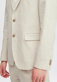 Beige linnet kavaj med notch-lapel, två framknappar, en bröstficka och knapppaneler på ärmsluten. Slät textur.