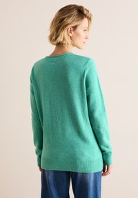 Ljus turkos ulltröja med ribbat halsringning och manschetter, med en avslappnad passform och något längre baktill. Texturerad ytfinish synlig.