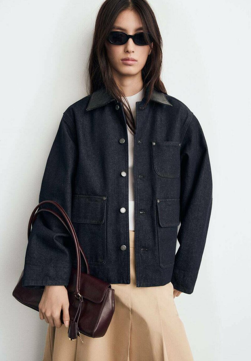 Dark denim jacket with a black collar, snap buttons, and front pockets, paired with a beige skirt and burgundy handbag. Black sunglasses worn.